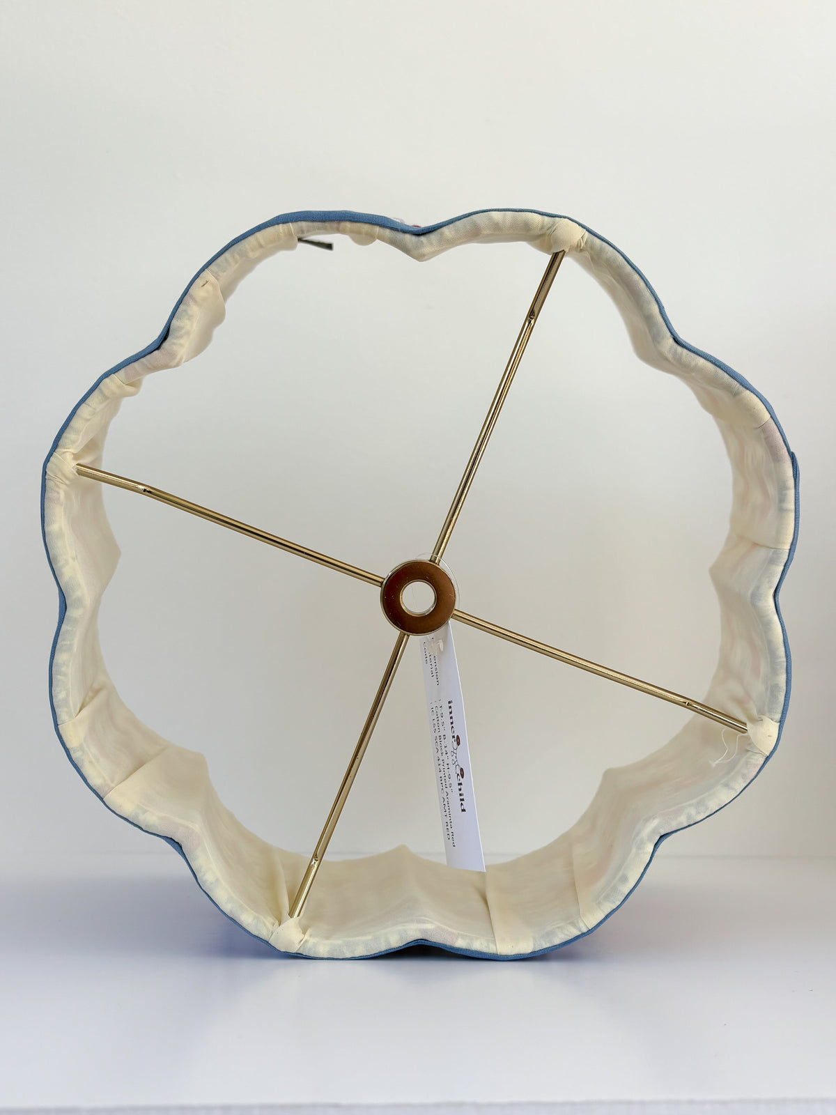 A scalloped flower-shaped fabric lampshade with a cream-colored inner lining, a blue outer edge, and gold-toned metal support rods, shown on a white surface against a white background.